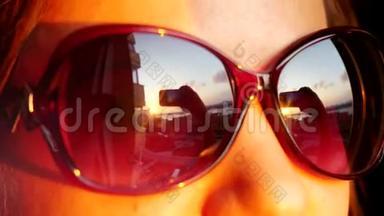 近景年轻女子戴着太阳镜`脸，站在窗前，拿着智能手机在日落时拍照。